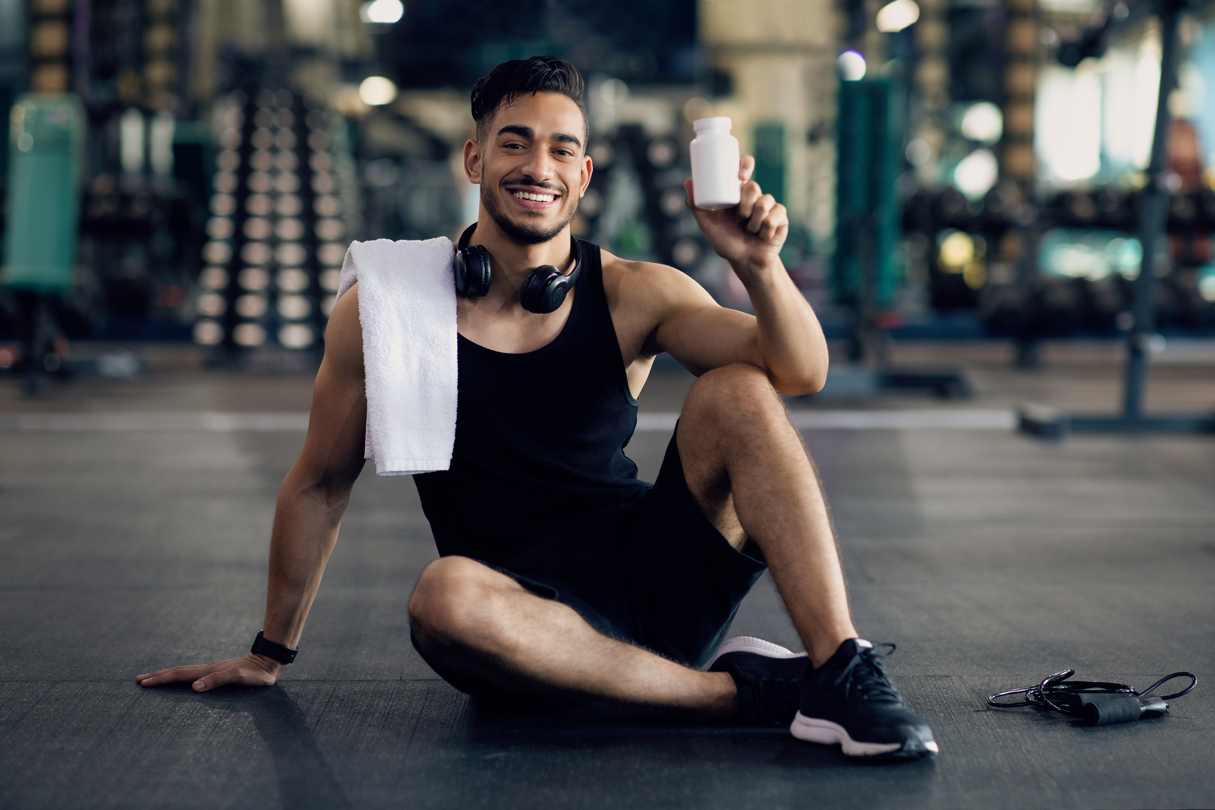 Young Muscular Arab Man Holding Container with Supplement Pills, Posing at Gym